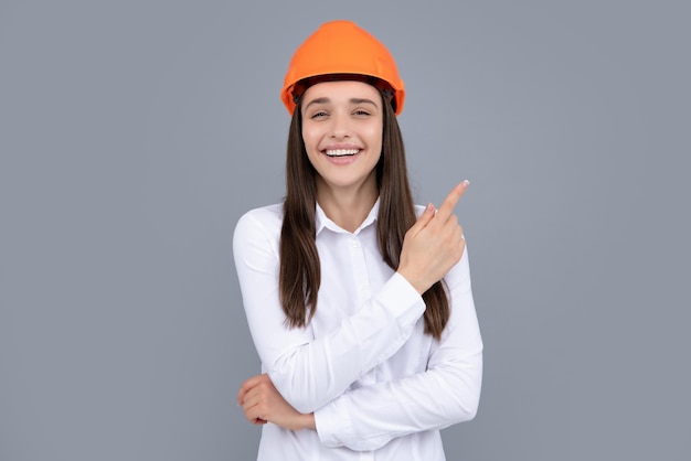 Young worker woman in helmet pointing on copy space on gray background