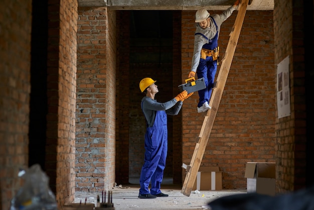 Young workman in blue overalls and hard hat giving toolbox to his colleague on ladder 