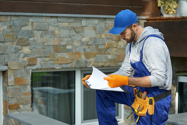 Young workman in uniform looking focused, standing outdoors with papers while working on construction project