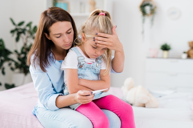 Young worried brunette female touching forehead of her sick daughter while looking at thermometer