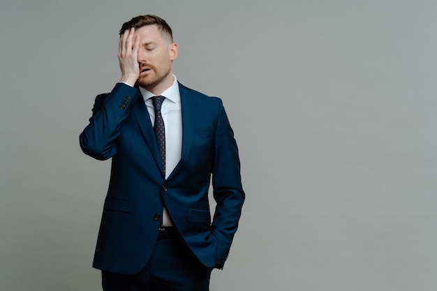 Photo young worried man manager in formal wear feeling frustrated and upset about business failure, holding hand on face and keeping eyes closed while standing against grey background, copy space for text