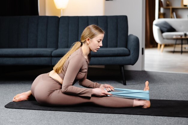 Молодая женщина-йог, практикующая йогу, делает упражнения, растягивая ноги с резинкой в спортивном бюстгальтере и брюках на полу в гостиной