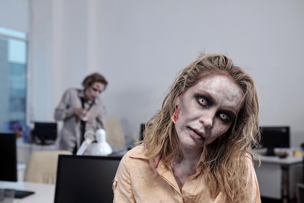Молодая женщина-зомби, стоящая перед камерой на фоне столов с компьютерными мониторами и мертвым коллегой-мужчиной