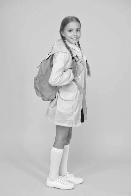 На спине и готов пойти в школу. Маленькая школьница носит милый плащ на желтом фоне. Очаровательный ребенок с школьным рюкзаком в дождливый день. Маленький ребенок снова в школу осенью.