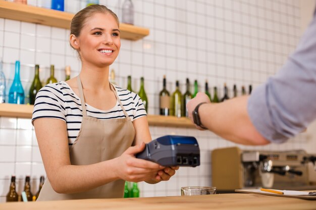 Your card please. Cheerful positive young woman holding a credit card terminal and looking at her client while asking for his card