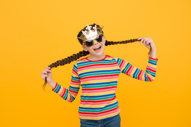 Your inner queen. happy princess hold hair yellow background. small child wear prop crown and glasses. party girl with long hair. kids barber. hair salon. haircare products. my hair my look