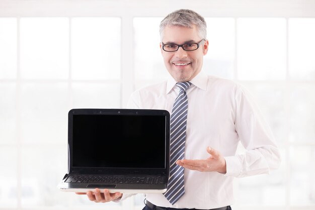 Il tuo testo qui. sorridente uomo anziano in camicia e cravatta che tiene il computer portatile e il monitor di puntamento