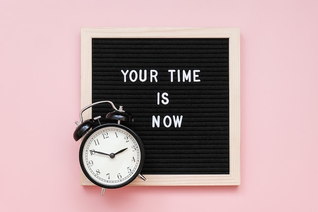 Your time is now. Motivational quote on letter board and black alarm clock on pink background