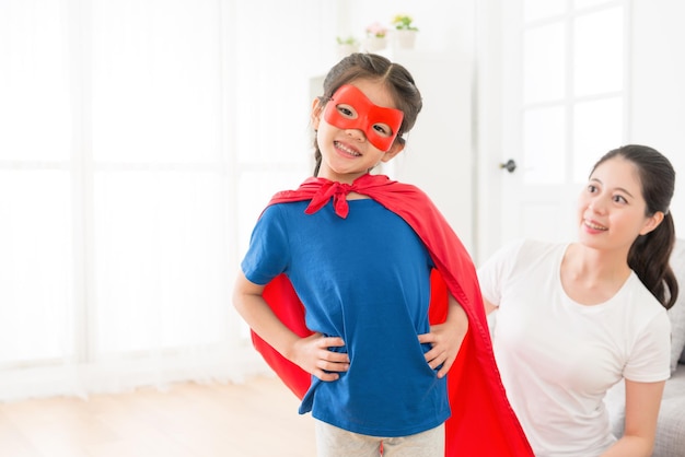 Photo youth beautiful little kid girl face to camera wearing red cloak with mask play as superhero and beauty smiling mother looking at her.