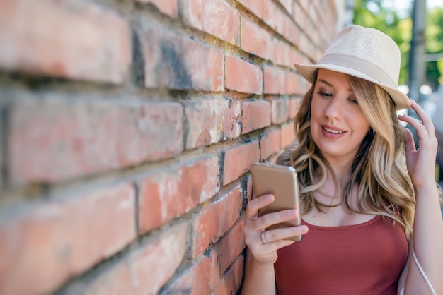 Photo youth and technology. attractive young woman in hat using smartphone and smiling while standing against brick wall.