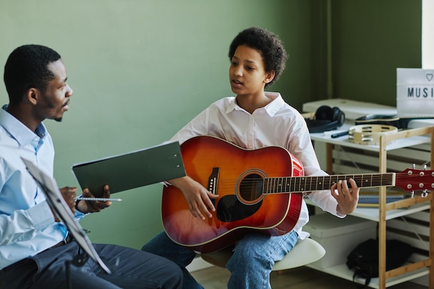Youthful african american schoolgirl playing acoustic guitar at lesson in music school while sitting