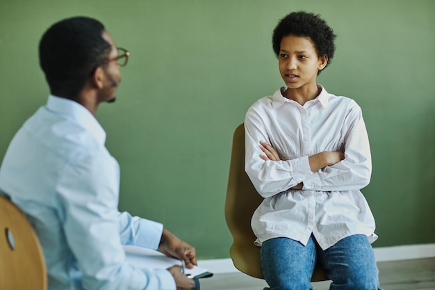 Youthful anxious schoolgirl describing her problem to counselor at psychological session while sitti