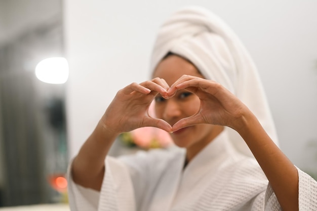 Photo youthful asian woman doing heart symbol shape with hands smiling to her reflection in mirror beauty and self care concept