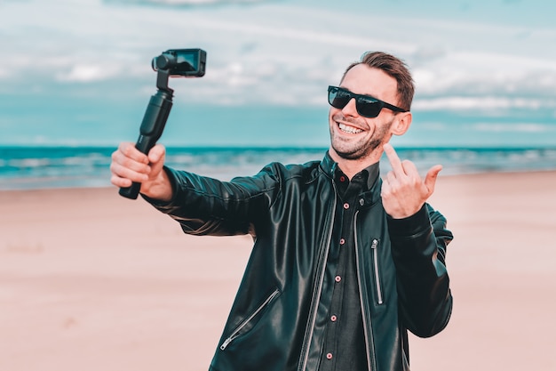 Gimbal Camera Stabilizer와 함께 액션 카메라를 사용하여 해변에서 셀카를 만들거나 비디오를 스트리밍하는 선글라스를 쓴 젊은 블로거.