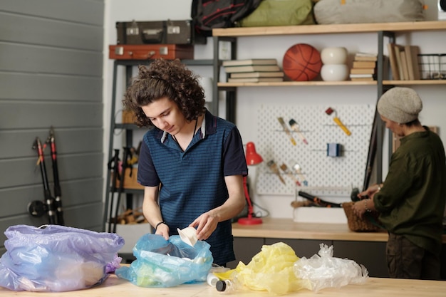 Youthful guy in casualwear putting plastic food containers into one of sacks