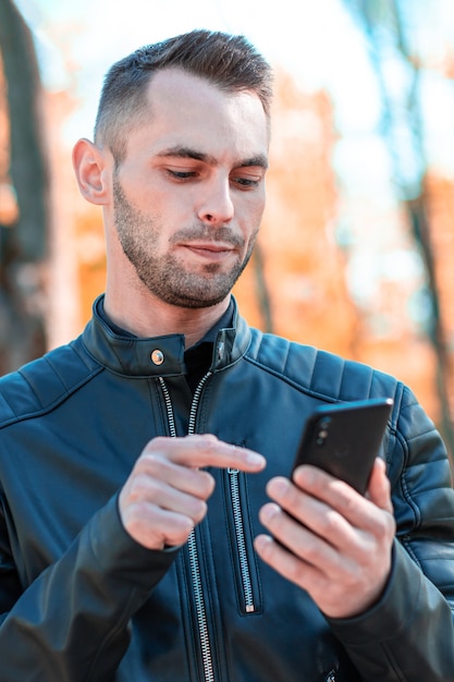 美しい秋の公園で黒いスマートフォンを使用して若い男。晴れた日に携帯電話を持つハンサムな若い男-中程度のクローズアップショット