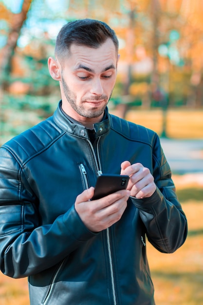 若い男は美しい秋の公園で黒いスマートフォンを使用して驚いた