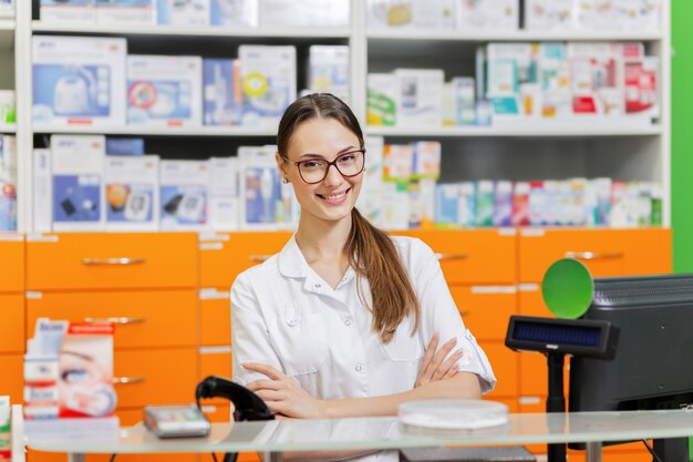 A youthful pleasant dark-haired girl with glasses,dressed in a medical overall,greets visitors at the cash desk in a new pharmacy. .