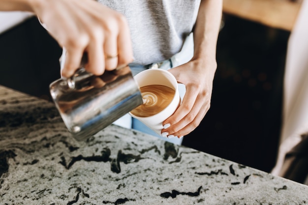 A youthful slim kind blonde girl,dressed in casual outfit,skillfully adds milk to coffee in a modern coffee shop. .