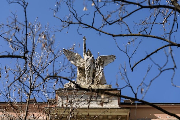 Zagreb Croatia March 28 2019 White angel statue on building roof view through tree brunches against blue sky Winged women with her right arm raised stone sculpture detailed view