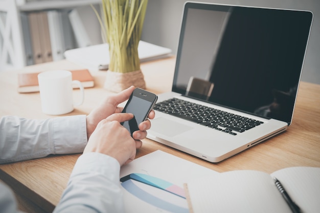 Zakelijk bericht aan het typen. Close-up van een man die een mobiele telefoon vasthoudt terwijl hij aan het houten bureau zit met een laptop erop