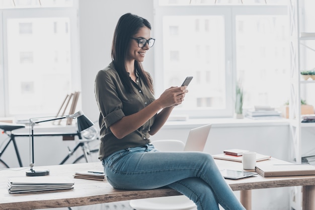 Zakelijk bericht aan het typen. Zelfverzekerde jonge vrouw in slimme vrijetijdskleding die een smartphone vasthoudt en ernaar kijkt met een glimlach terwijl ze in de buurt van haar werkplek staat