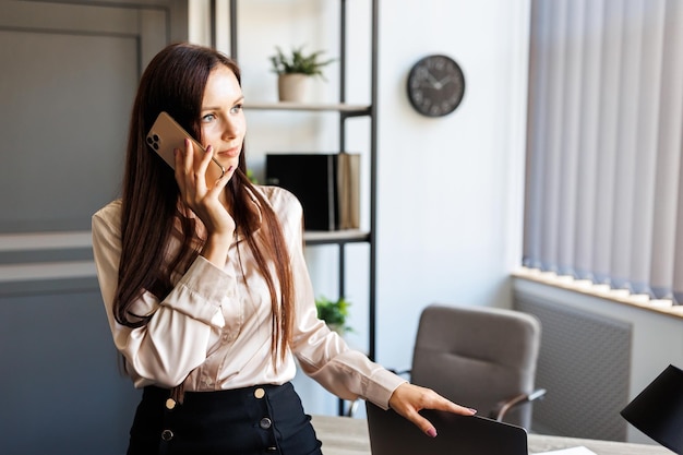 Zakelijk gesprek Vrolijke jonge mooie vrouw die op haar mobiele telefoon praat en naar het raam kijkt terwijl ze op de tafel op haar werkplek leunt