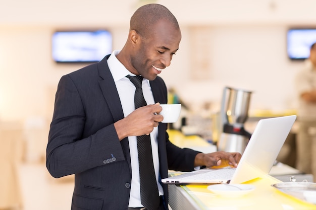 Zakelijk onderweg. Vrolijke jonge Afrikaanse man in formalwear die zijn laptop gebruikt terwijl hij aan de bar staat