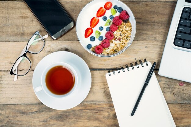 Zakelijk ontbijt met een kom yoghurt, een kop thee, een mobiele telefoon, een laptop, een computer en een notitieblok op de houten tafel.