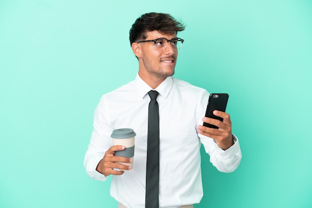 Zakelijke blanke man geïsoleerd op blauwe achtergrond met koffie om mee te nemen en een mobiel terwijl hij iets denkt