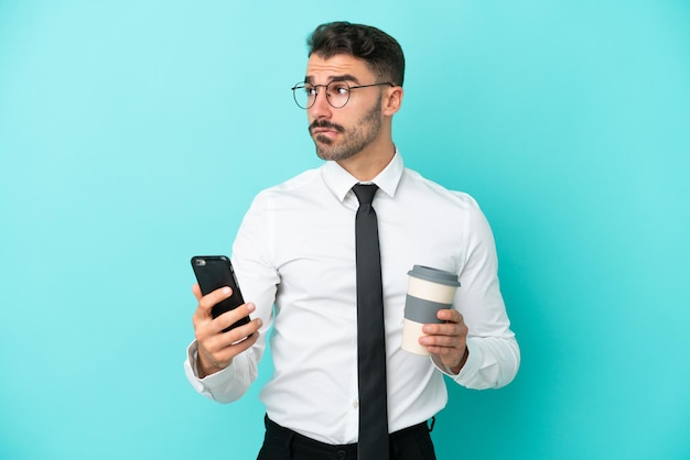 Zakelijke blanke man geïsoleerd op blauwe achtergrond met koffie om mee te nemen en een mobiel terwijl hij iets denkt