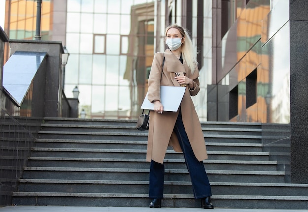 Zakelijke blonde vrouw met een medisch masker houdt een laptop in haar handen op de achtergrond van de trap