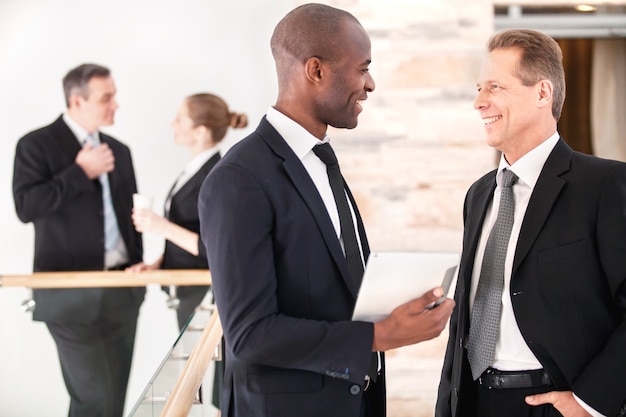 Zakelijke communicatie. Twee vrolijke zakenmannen die met elkaar praten terwijl hun collega's op de achtergrond staan