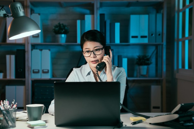 zakelijke deadline en technologieconcept. Aziatische Japanse zakenvrouw met laptopcomputer die 's nachts op kantoor belt. vrouwelijke werknemer die discussie heeft met collega's tijdens overuren.