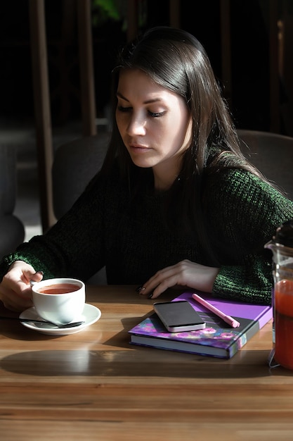 Zakelijke jonge brunette zit aan een tafel in een café en drinkt thee voor haar telefoon