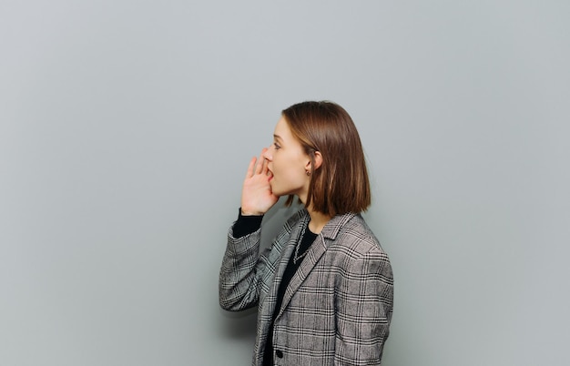 Foto zakelijke jonge vrouw in een jas schreeuwt in de handpalmen opzij op kopieerruimte op een grijze achtergrond