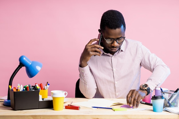 Zakelijke ongeschoren Afro-Amerikaanse man met bril, gestreept shirt, praten met zakenpartner op smartphone, zittend aan een bureau omringd met briefpapier, geïsoleerd op roze muur.