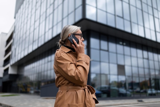 Zakelijke senior volwassen vrouw in een herfstjas op de achtergrond van de glazen gevel van het kantoor