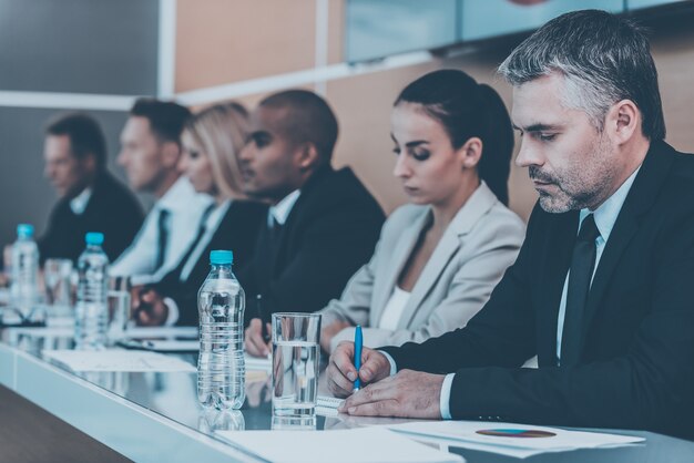 Zakelijke vergadering. Zijaanzicht van zakenmensen die iets in hun notitieblokken schrijven terwijl ze op een rij zitten