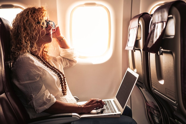 Zakelijke volwassen mooie krullende blonde vrouw reist op vliegtuig verbonden met internet met moderne computerlaptop