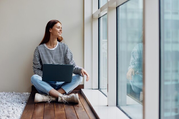 Zakelijke vrouw zit met een laptop bij het raam vol lengte en glimlachende gelukkige vrouw werkt online in de kantoor kopie ruimte