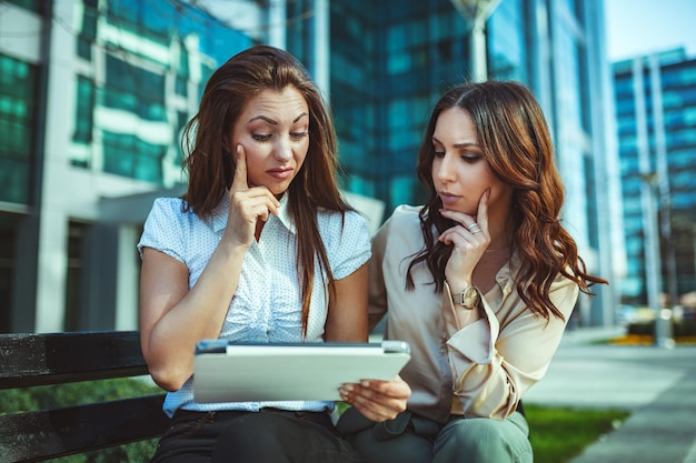 Zakelijke vrouwen zitten in het kantoorpark terwijl ze een digitale tablet gebruiken en een wegwerpkoffiekopje vasthouden. Business team werkt online saamhorigheid tijdens het raadplegen.