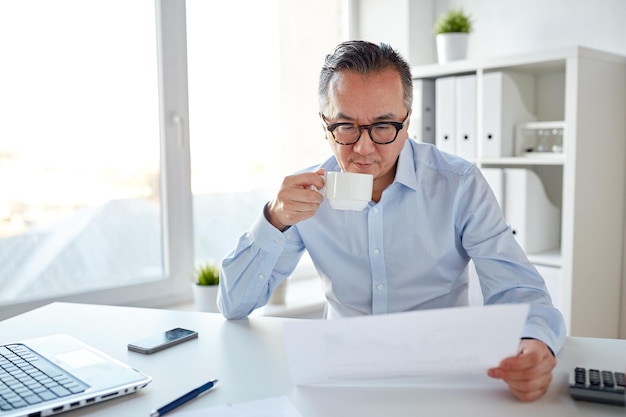 zaken, mensen, papierwerk en technologieconcept - zakenman die papier leest en koffie drinkt op kantoor