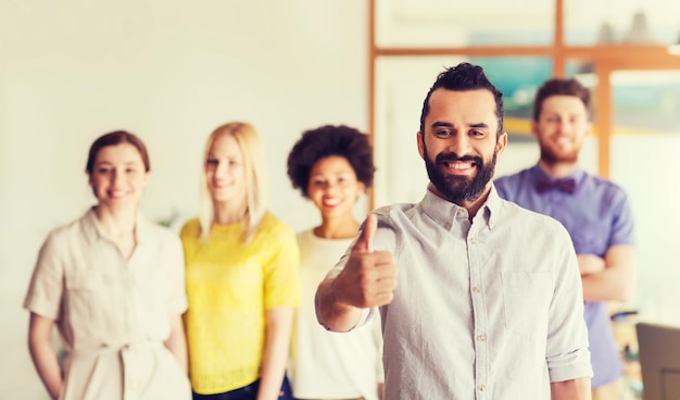 zaken, opstarten, mensen, gebaar en teamwork concept - gelukkige jonge man met baard duimen opdagen over creatief team in kantoor