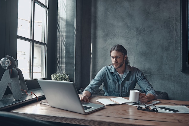 Zaken regelen. Nadenkende jonge man aan het werk met behulp van de computer en een kopje te houden