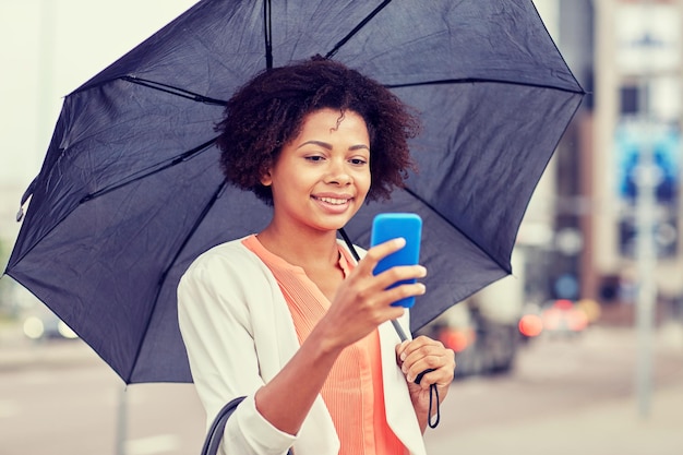 zaken, slecht weer en mensen en concept - gelukkige Afro-Amerikaanse zakenvrouw met paraplu sms'en op smartphone in city street