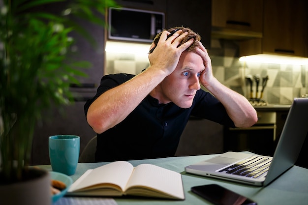 Zakenman aan de tafel thuis, modern gezellig huis, kijkend naar laptopscherm, voelt zich ontevreden over het verrichte werk, succesvolle man is nerveus en steekt zijn handen achter zijn hoofd