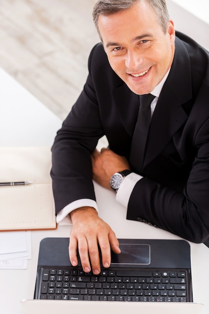 Zakenman aan het werk. Bovenaanzicht van een gelukkige volwassen man in formalwear die op laptop werkt en glimlacht terwijl hij op zijn werkplek zit