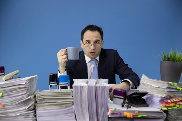 Zakenman boos kijken naar de voorkant met een kopje koffie in zijn hand aan een bureau vol papierwerk