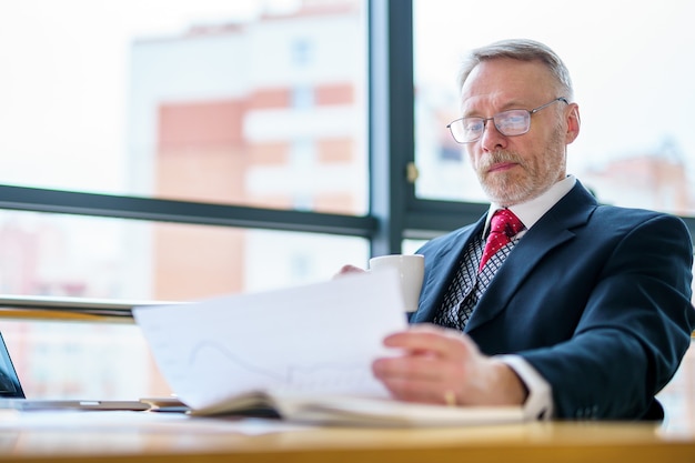 Zakenman controleert een afgedrukt rapport of document terwijl hij aan tafel zit en werkt aan papierwerk op een laptop om zijn standpunt uit te leggen. Foto in een kantoor.